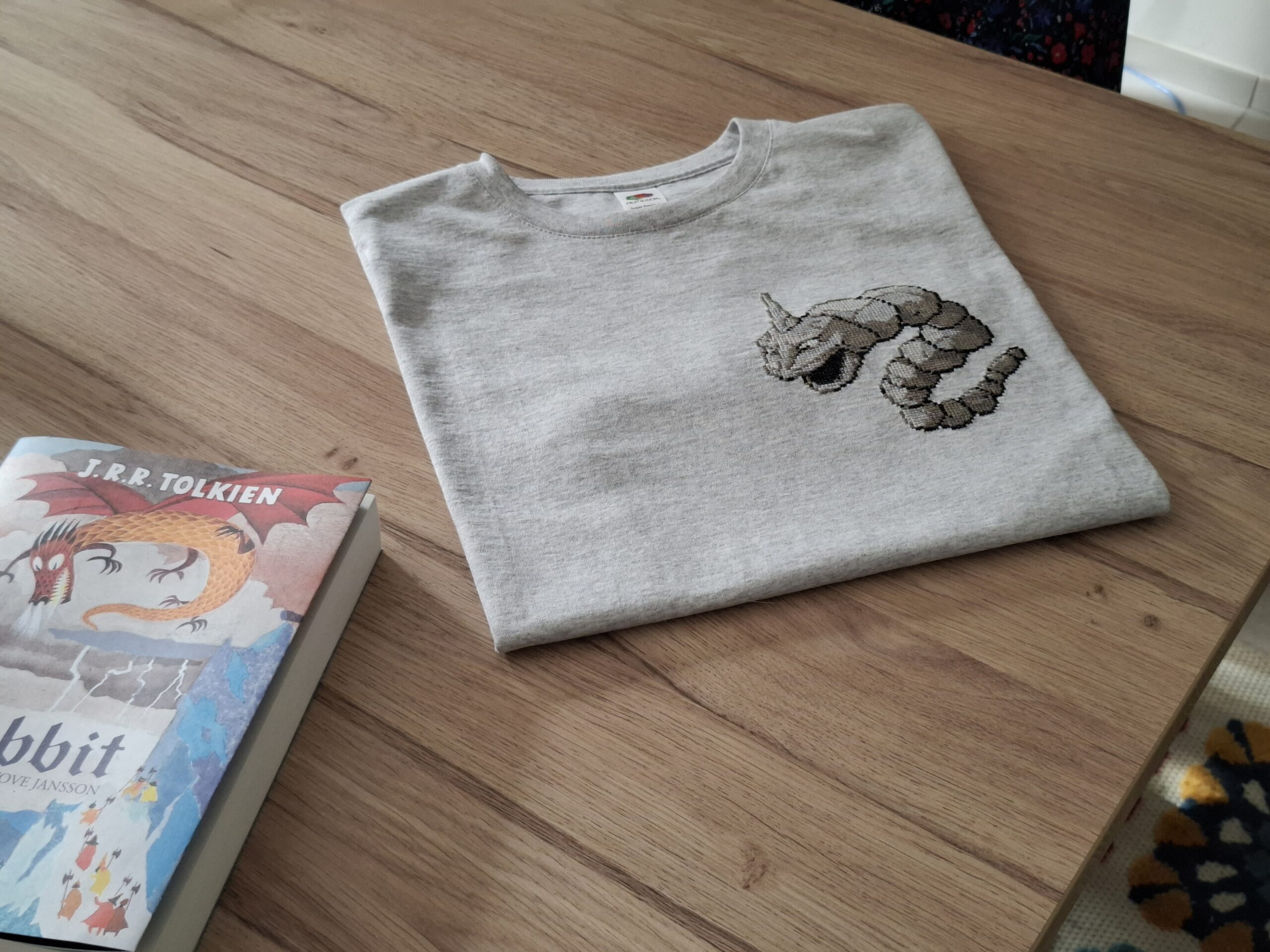 T-shirt onix plié, posé sur une table en bois accompagné d'une édition du Hobbit illustrée par Tove Jansson.